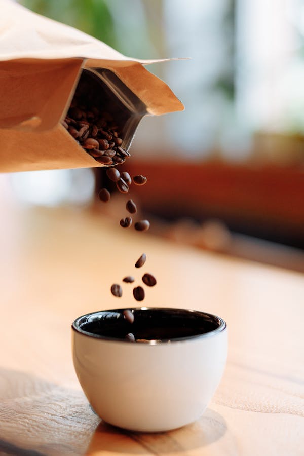 Coffee beans being poured from a paper bag into a white cup on a wooden table.