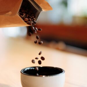 Coffee beans being poured from a paper bag into a white cup on a wooden table.