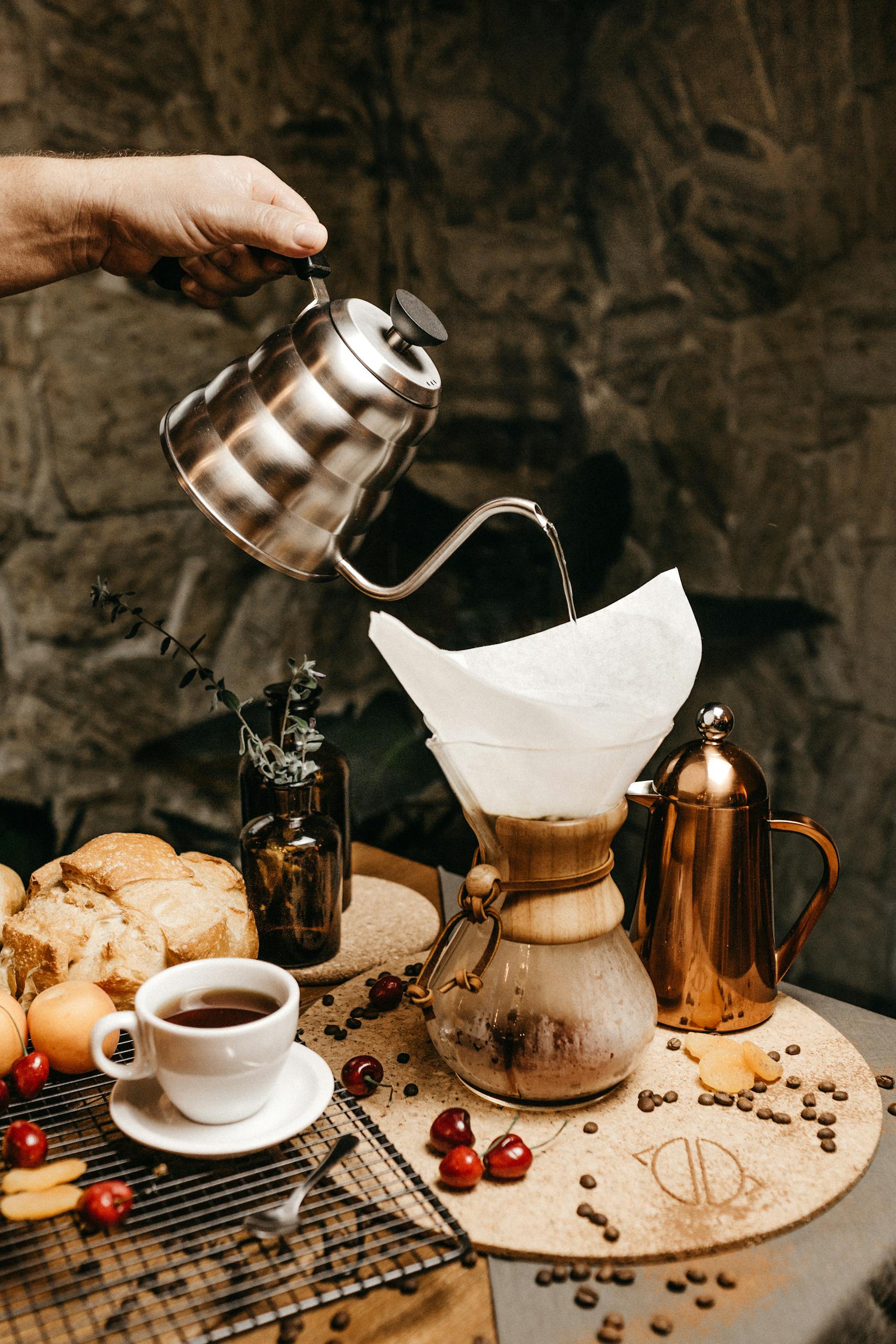 Hand pouring coffee into a Chemex surrounded by fresh cherries and breakfast items indoors.
